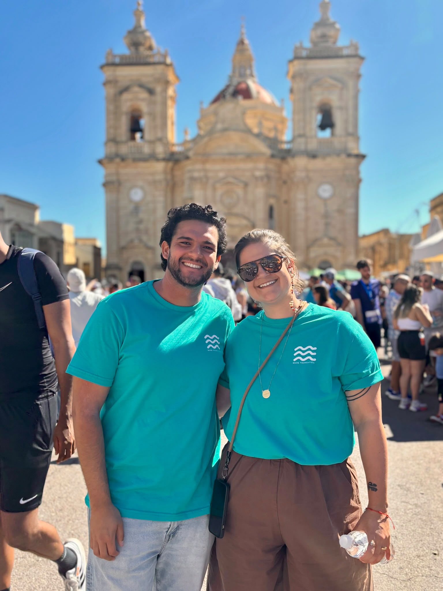 Two MARE Foundation volunteers in branded turquoise t-shirts at the RunGozo event, with a Gozitan church behind the crowd.